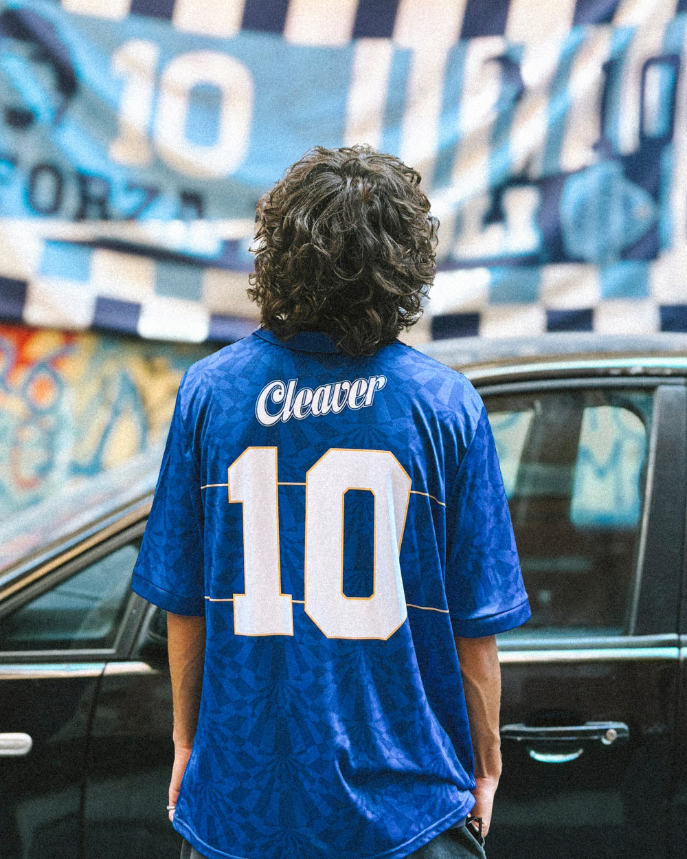 Person wearing a blue jersey with 'Cleaver' and number '10' in front of a car and wall with graffiti.